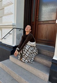 Egy barna felsőt és kockás szoknyát viselő nő ül egy épület előtt, ahol egy fából készült ajtó található, és kezét összekulcsolva néz felfelé.