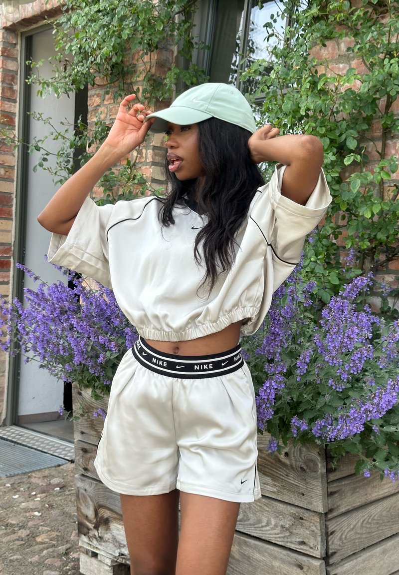 Femme en tenue de sport claire et casquette vert pâle ajustant son chapeau, debout à côté d'une jardinière en bois avec des fleurs violettes et un feuillage vert.