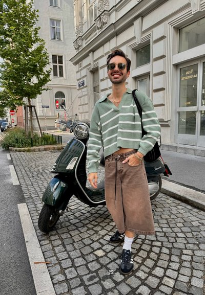 Suéter corto a rayas verde y blanco, pantalones cortos de pierna ancha marrones, zapatillas negras con calcetines blancos, de pie al lado de un patinete verde sobre adoquines.