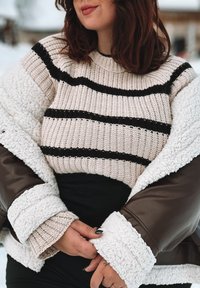 Maglione a righe beige con linee nere e bianche, indossato sotto un giubbotto in pelle marrone foderato in pile bianco, con dettagli in tessuto testurizzato visibili.