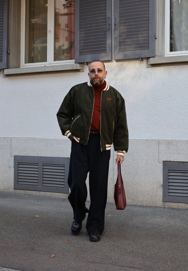 Un hombre con gafas de sol camina por la acera con un bolso de cuero color burdeos, vestido con una cazadora bomber oscura y unos pantalones negros holgados.