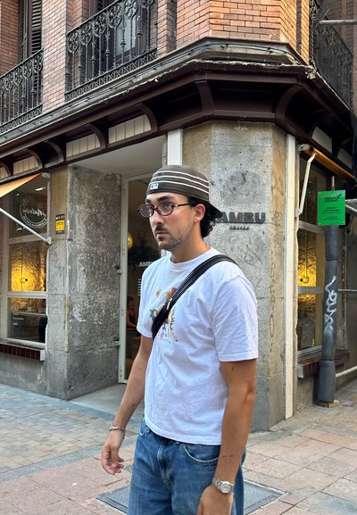 Hombre vestido con una camiseta gráfica blanca y jeans azules, con gafas y una gorra a rayas, de pie al aire libre cerca de un edificio de piedra.