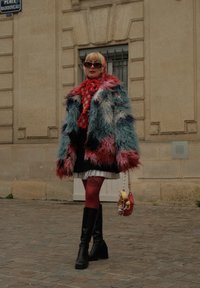 Kolorowy płaszcz z futerka ekologicznego w odcieniach niebieskiego, różowego i białego, czerwona kraciasta szal, czarna spódnica, czerwone rajstopy i czarne kozaki do kolan. Mała torebka.