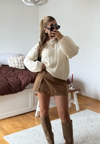 Maglione bianco a trecce, gonna mini marrone con pieghe laterali, stivali marroni al ginocchio, collane d'oro a più strati, occhiali da sole neri, sfondo neutro.