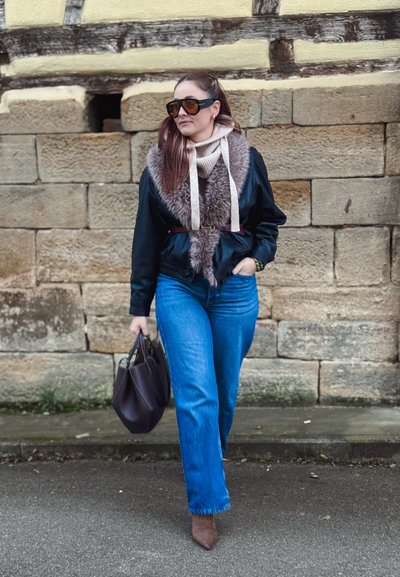 Chaqueta de cuero negra con cuello de piel, suéter crema con lazos, vaqueros azules, botas marrones con tacón y un bolso oscuro. Fondo de muro de piedra.