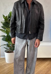 Veste en cuir noir avec fermeture éclair à l'avant, col large et détails cousus; portée avec un pantalon ample gris clair.