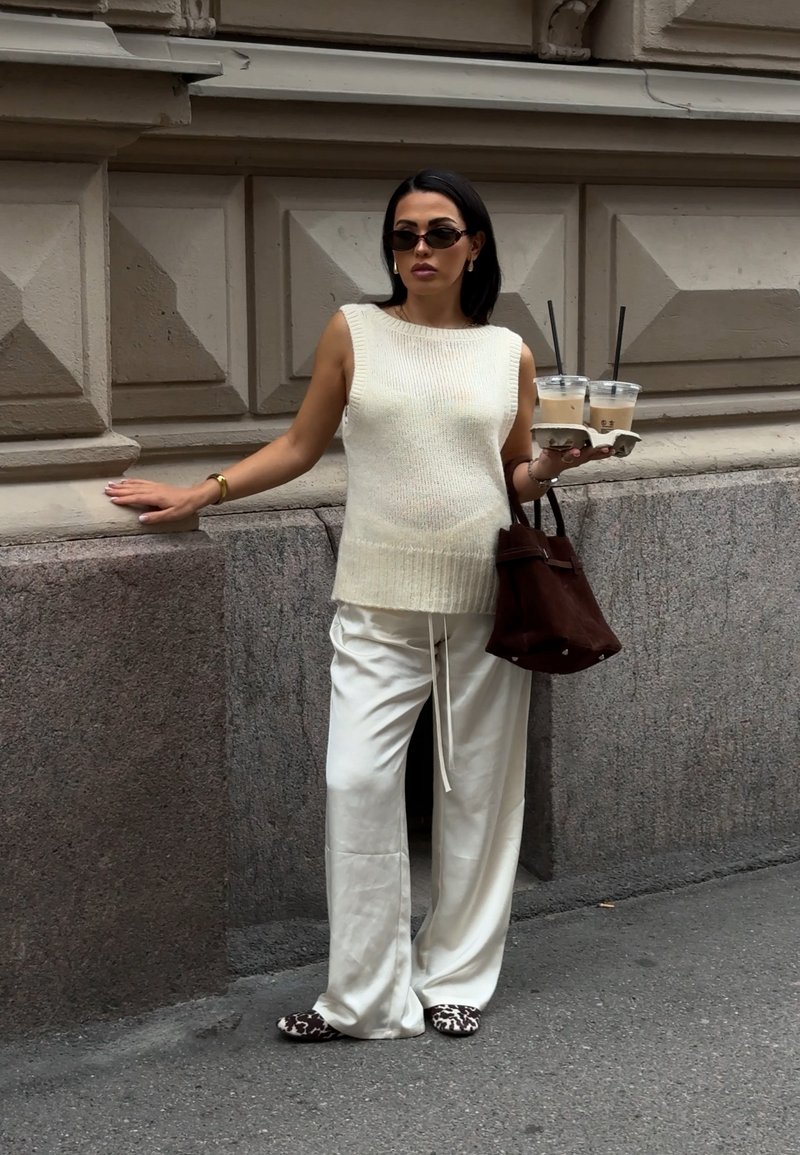 Top sans manches en tricot blanc, pantalon ajusté crème à larges jambes, sac en suède marron, tenant deux cafés glacés. Lunettes de soleil noires, pantoufles à imprimé léopard.