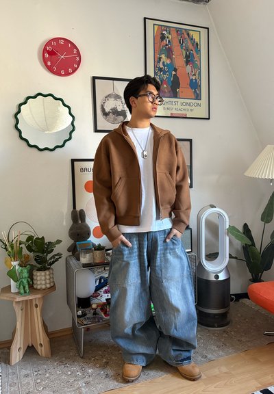 Joven con chaqueta marrón y vaqueros holgados está de pie en una habitación decorada con plantas, ventilador, reloj y arte enmarcado en paredes blancas.