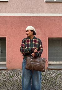 Femme en jean large et veste à carreaux tenant un sac en cuir marron, portant un béret blanc et des boucles d'oreilles dorées, debout près d'un mur rose texturé.