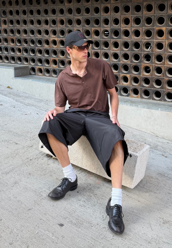Homme portant une casquette noire, des lunettes de soleil, une chemise brune, un short noir et des chaussures noires assis sur un bloc de béton près d’un mur avec des ouvertures circulaires.