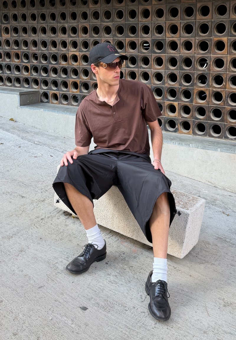 Homme portant une casquette noire, des lunettes de soleil, une chemise brune, un short noir et des chaussures noires, assis sur un bloc de béton près d'un mur avec des ouvertures circulaires.