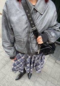 Veste bomber grise à coupe oversize, associée à une jupe à carreaux. Sac à main noir orné d'un pendentif en forme de croix. Bottines noires pointues complètent le look.