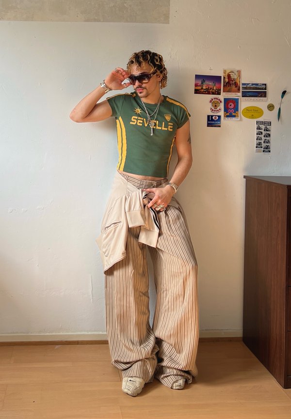 Persoană purtând un tricou verde scurt „Seville”, pantaloni largi bej cu dungi, ochelari de soare și bijuterii, stând lângă un perete cu autocolante de călătorie.