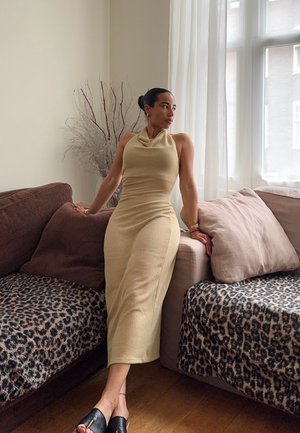 Knit, sleeveless, beige dress with a cowl neckline. Set against a brown couch and a leopard print throw. Black slide sandals. Natural light.