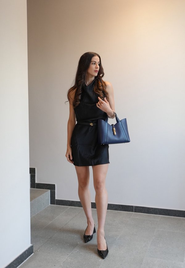 Mujer con vestido negro sin mangas y tacones negros que sostiene un bolso azul marino, está de pie en el interior cerca de unas escaleras, mirando hacia su izquierda.