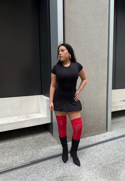 Vestido negro ajustado con dobladillo acampanado, combinado con medias rojas altas hasta el muslo con detalles de encaje y botas de tacón alto negras.