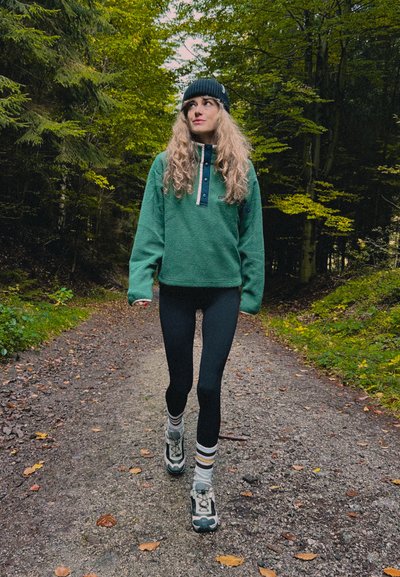 Sudadera de forro polar verde con tapeta de botones azul, leggings negros y zapatillas grises con calcetines a rayas multicolor, caminando por un sendero del bosque.