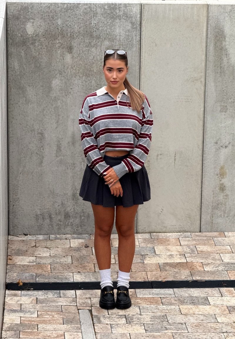 Jeune femme avec des lunettes sur la tête, portant un pull rayé court, une jupe plissée noire, des chaussettes blanches et des mocassins noirs, debout sur un sol carrelé.