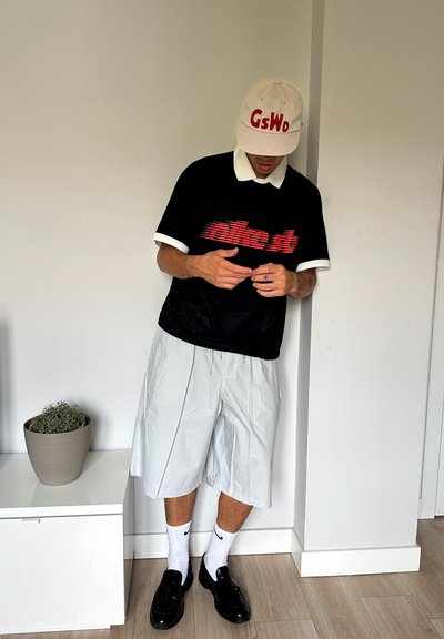 Camisa negra de manga corta con el texto rojo "nike.st", cuello blanco, pantalones cortos gris claro, calcetines blancos de Nike y mocasines negros. Planta en una maceta cerca.