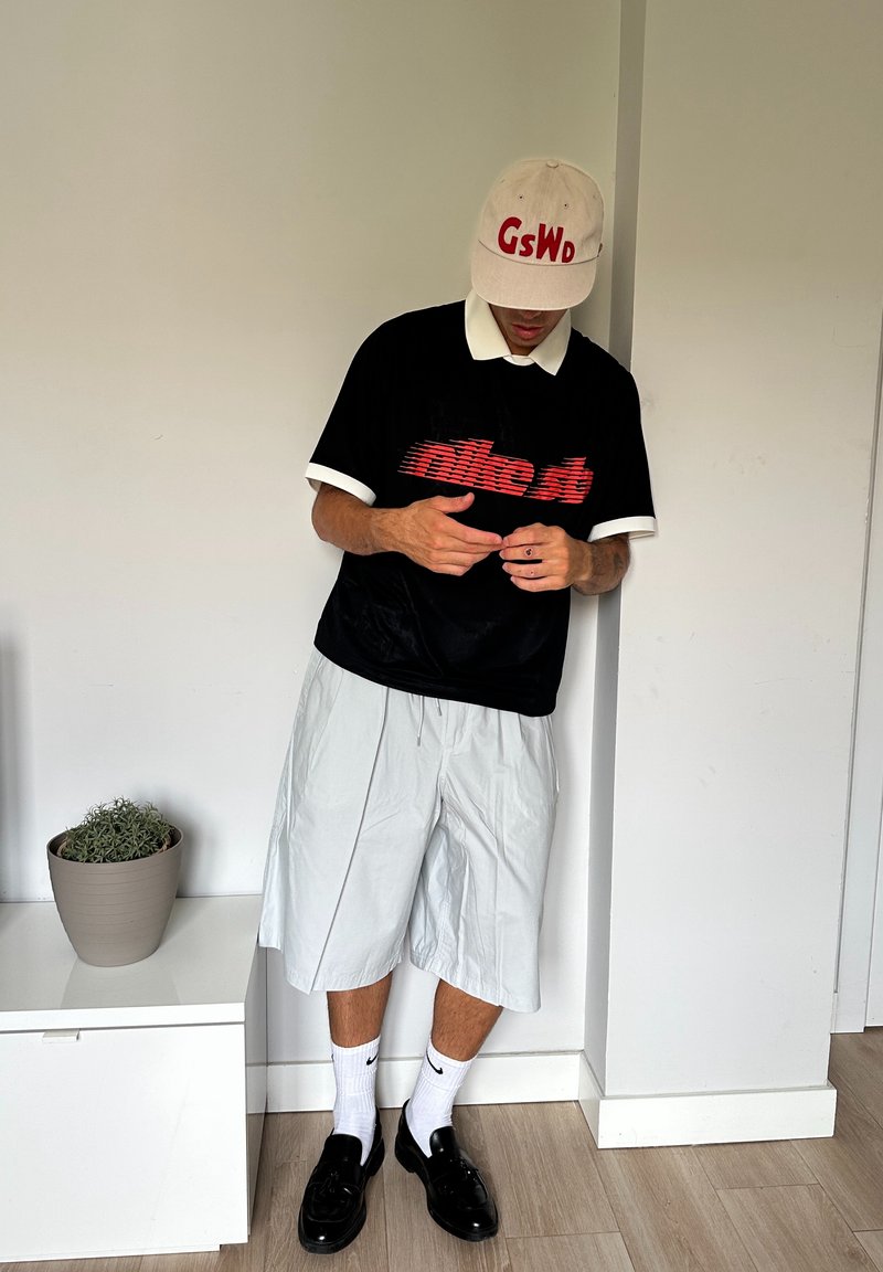 Chemise noire à manches courtes avec le texte rouge "nike.st", col blanc, shorts gris clair, chaussettes blanches Nike et mocassins noirs. Plante en pot à proximité.