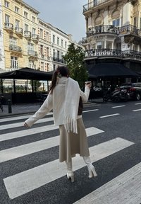 Suéter blanco de punto con patrones de trenzas, superpuesto sobre una falda midi beige. Tacones blancos de tiras y un bolso marrón. Taza de café en la mano.