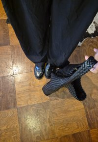 Person wearing black trousers and black leather loafers holding a woven black handbag over a wooden parquet floor.