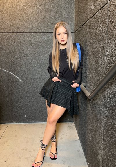 Top negro de manga larga, falda negra de rayas con vuelo, tacones negros con tiras y tachuelas, y un bolso azul de hombro. Cabello liso y recto.
