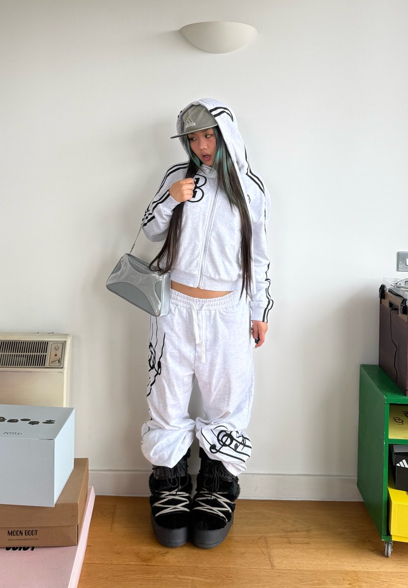 Personne portant un sweat à capuche blanc et un pantalon assorti avec des motifs noirs, une casquette Adidas grise, un sac à main argenté et des bottes noires doublées de fourrure, debout à l'intérieur.