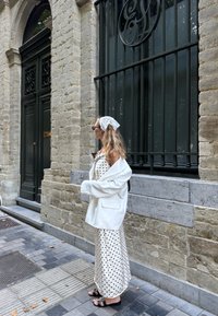 Blazer oversized bianco sopra un abito maxi a pois. L'abito è beige con pois neri. Occhiali da sole scuri e una sciarpa bianca completano il look.