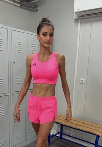 Ružový športový crop top a šortky s logom, vyrobené z ľahkého materiálu. Pozadie zahŕňa skrinky a lavičku.