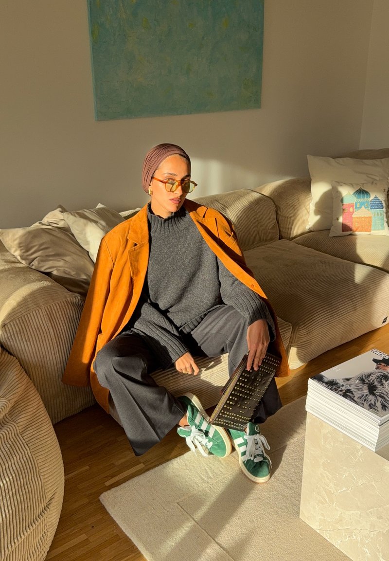 Manteau en daim orange, pull gris, pantalon noir, baskets vertes à rayures blanches. Assis sur un canapé rayé, tenant un objet noir texturé.