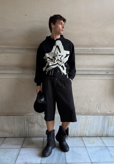 Sudadera negra con un gran gráfico de estrella blanca, combinada con pantalones cortos negros holgados y botas negras forradas de piel. Sosteniendo un sombrero de piel negro.