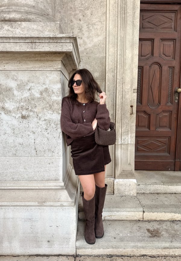 Kobieta w ciemnobrązowym kardiganie, spódnicy i butach stoi obok kamiennego budynku z dużymi drewnianymi drzwiami, trzymając plecioną torebkę i okulary przeciwsłoneczne.