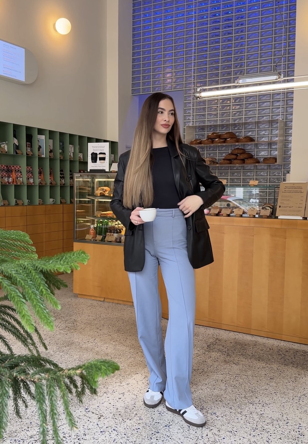 Young woman in a black jacket, blue trousers, and trainers holding a coffee cup, standing inside a bakery with bread and pastries on display.