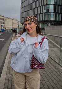 Sweatshirt cinza claro com fecho éclair e padrão texturizado, calças cargo bege e uma mala bordeaux decorada com tachas prateadas. Faixa de cabelo com padrão de estampado de leopardo.