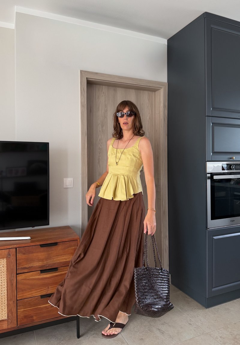 Donna con occhiali da sole, top giallo senza maniche, gonna lunga marrone e sandali, che tiene una borsa nera intrecciata, in piedi in una cucina-soggiorno moderna.