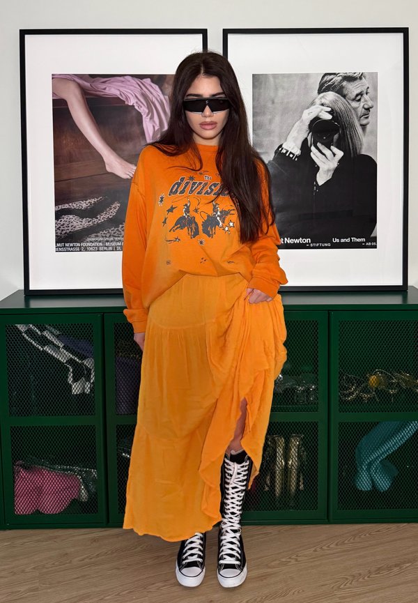 Femme aux longs cheveux foncés portant des lunettes de soleil noires, un sweat-shirt graphique orange, une jupe assortie et des baskets montantes noires et blanches, debout à l'intérieur.