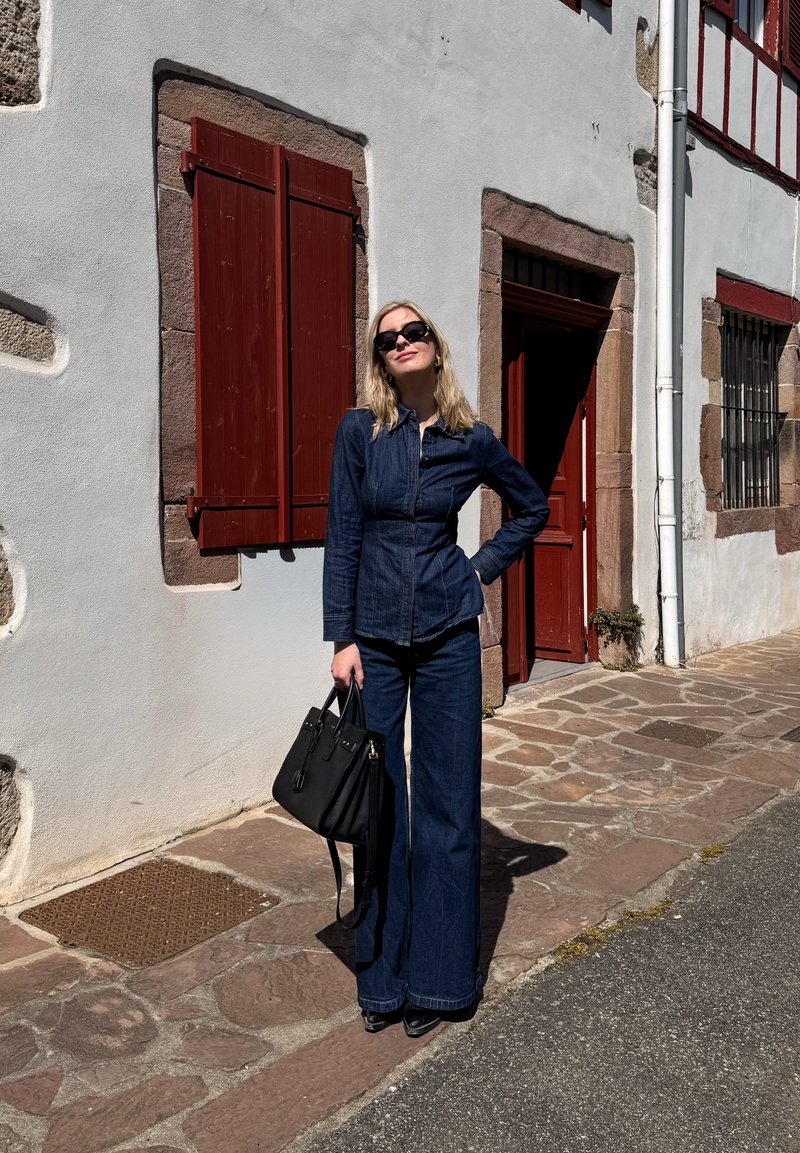 Mujer con conjunto de mezclilla oscuro y gafas de sol, sosteniendo un bolso negro, está de pie en una calle adoquinada junto a un edificio blanco con contraventanas y puerta rojas.