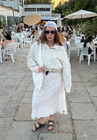 Fehér ruházat fekete pöttyökkel; gombos ing és hosszú szoknya. Kiegészítők közé tartozik egy fehér táska és sötét napszemüveg. Barna szandál csatokkal.