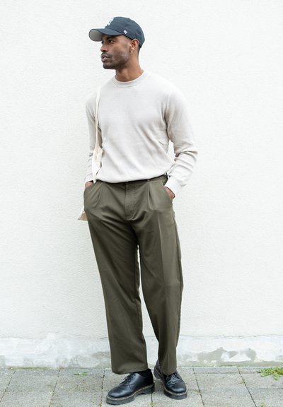 Suéter beige de canalé, pantalones de pinzas verde oliva, zapatos de cuero negros y una gorra gris. Diseño sencillo, ajuste casual, con un bolso de hombro.