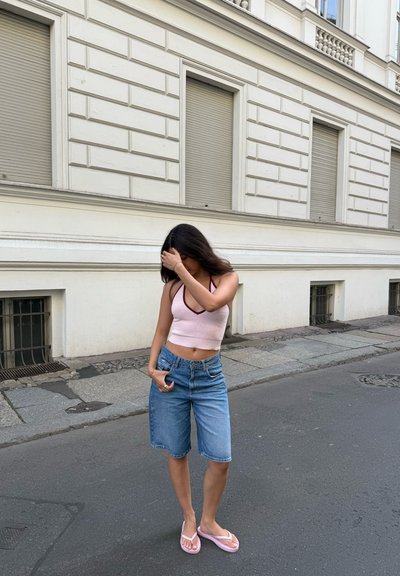 Top corto rosa claro con escote en V y ribete burdeos, combinado con pantalones cortos bermuda de mezclilla azul y chanclas rosas, de pie en una calle.
