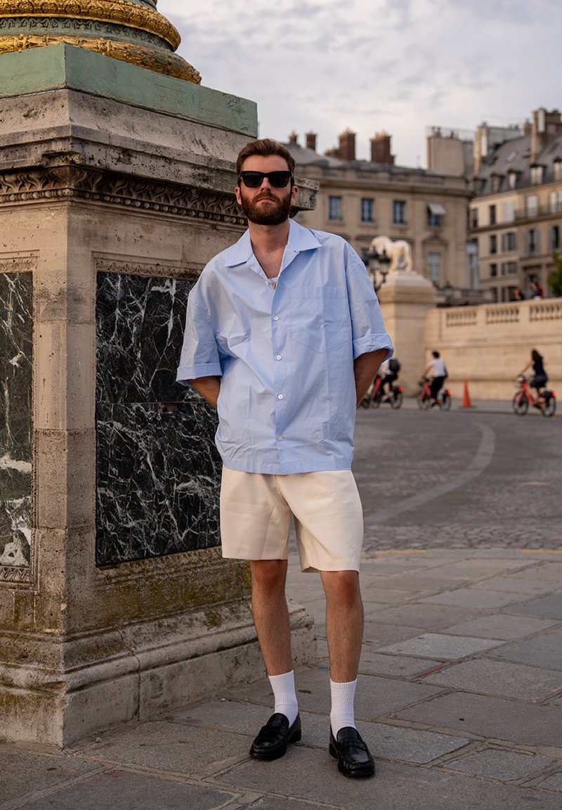 Homme barbu portant des lunettes de soleil, chemise à manches courtes bleu clair, short blanc, chaussettes blanches et mocassins noirs se tient près d'un monument en pierre.