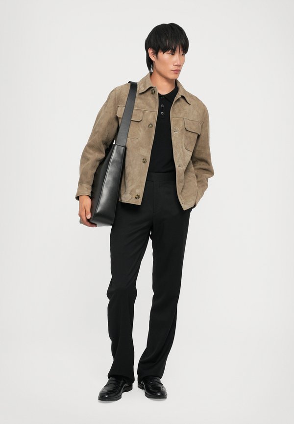Casaco de camurça beige com dois bolsos à frente, t-shirt polo preta, calças de fato pretas e sapatos pretos. Segura uma mala de ombro preta texturizada.