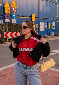 Tricou PUMA roșu și negru cu un model marmorat, asortat cu o geacă neagră, blugi bleu și o geantă galbenă.