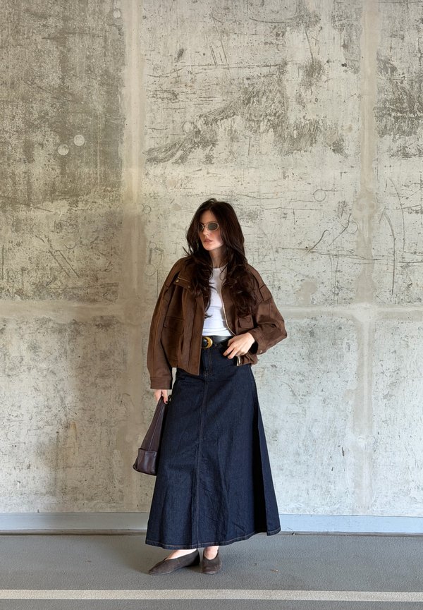 Mujer con pelo largo que lleva gafas de sol, chaqueta marrón, camiseta blanca, falda larga oscura, sosteniendo un bolso oscuro contra una pared de hormigón texturizada.