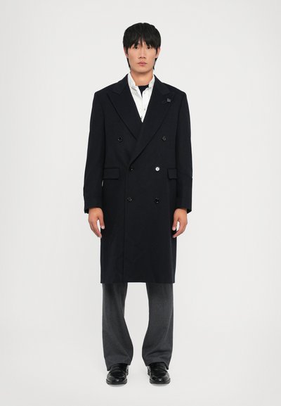 Abrigo de lana negro de doble botonadura con grandes botones, solapas con muescas y dos bolsillos frontales, combinado con pantalones grises y zapatos negros.