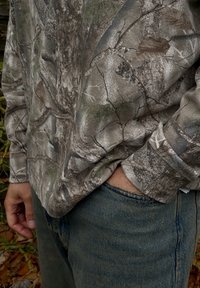 Camisa de manga larga con un patrón de camuflaje en tonos de gris, marrón y verde. Combinada con unos vaqueros azules, que muestran un corte relajado.