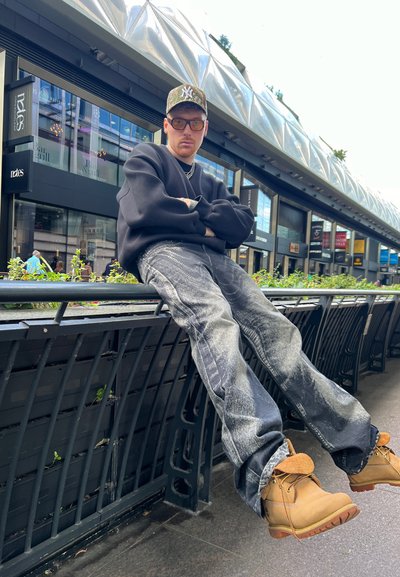 Persona con un sudadera negra de gran tamaño, jeans grises con estampado y botas de trabajo marrones, sentado en una barandilla negra con un fondo urbano.