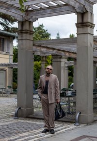 Μπεζ, υφαντό κοστούμι με μαύρο τοπ, φορεμένο με μαύρα παπούτσια. Το άτομο κρατά μια μαύρη τσάντα και στέκεται κάτω από μια ξύλινη περσίδα.