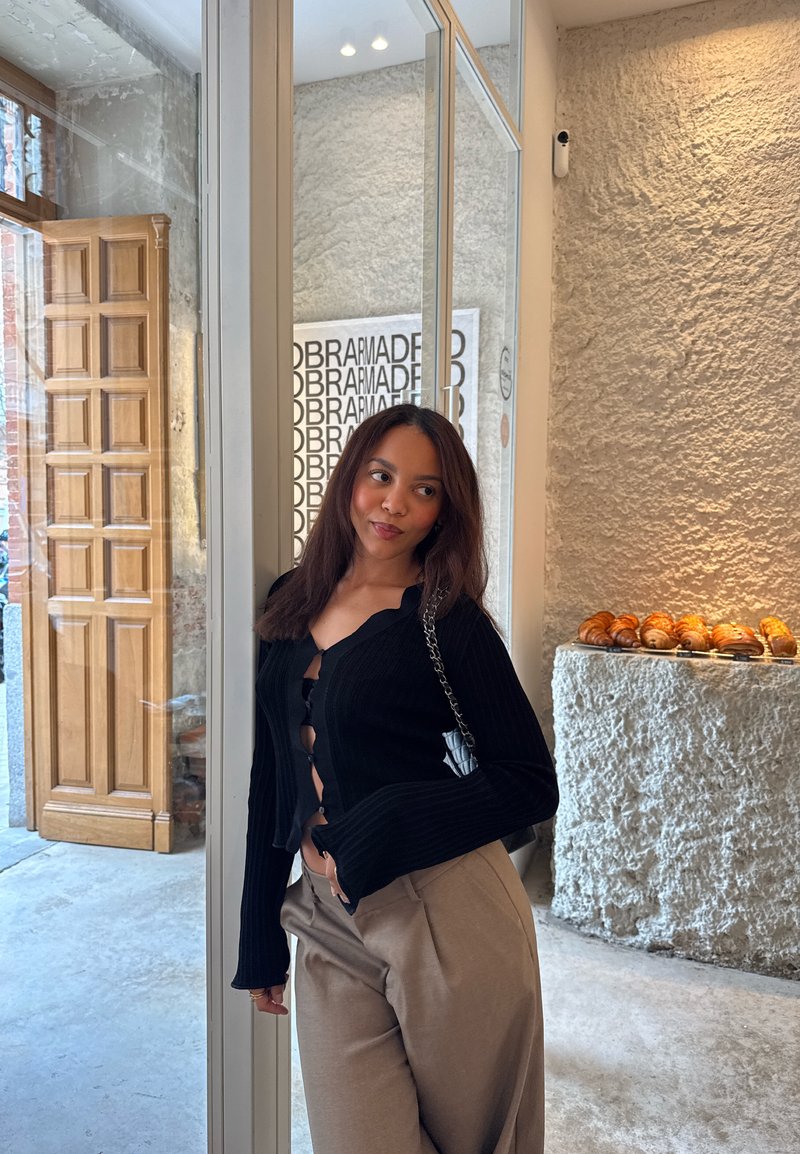 Giovane donna con top nero e pantaloni beige appoggiata a una porta di vetro all'interno di un caffè con pareti in pietra e pasticcini in esposizione.